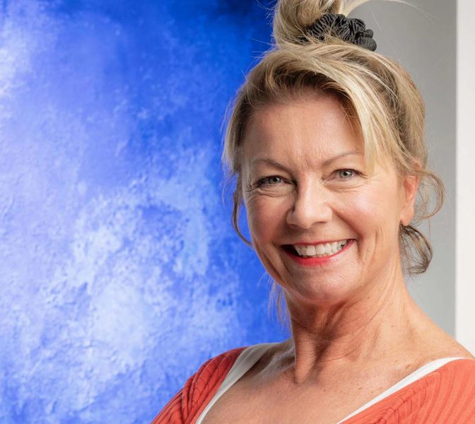Smiling woman in front of a blue abstract painting, wearing a red top with hair tied up.