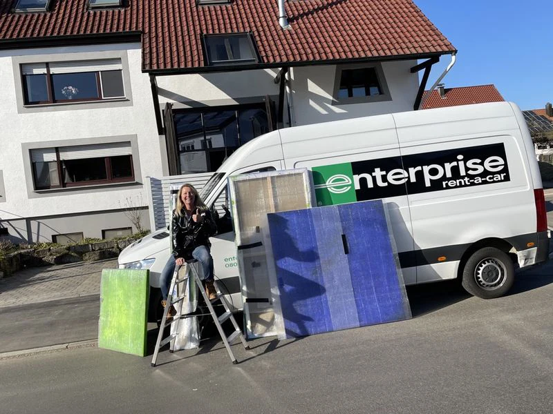 Woman sitting on a ladder beside abstract paintings and a rental van from Enterprise.