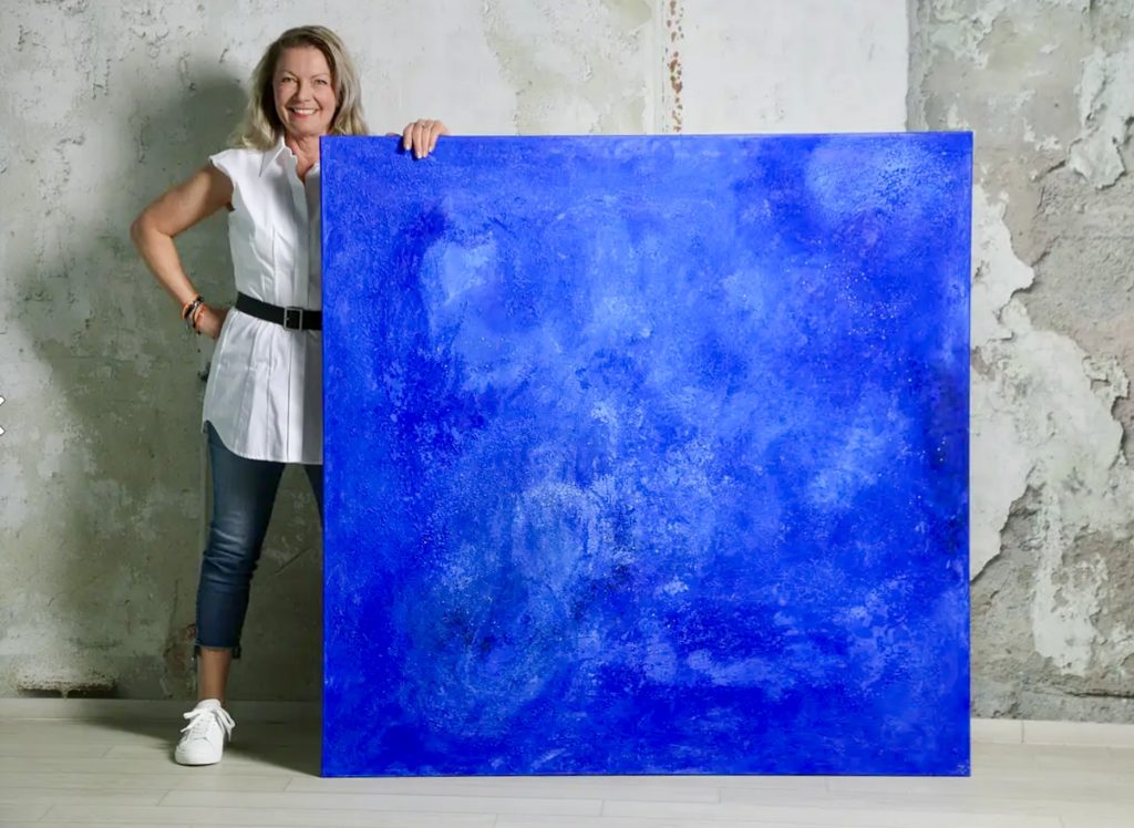 Woman standing beside a large blue abstract painting against a textured wall.