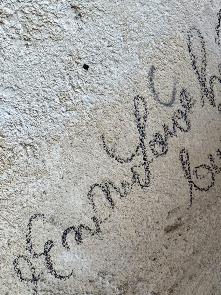 Detailaufnahme einer hellen Wand oder Putzoberfläche mit handschriftlichen Notizen oder Kritzeleien.